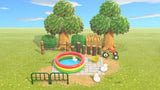 Animal Crossing Duckie Splash Pad room design with a colourful plastic pool, toy ducks, a yellow bath bucket and a red tricycle on stone paving, enclosed by barbed wire fencing, log stakes, hedges and hardwood trees